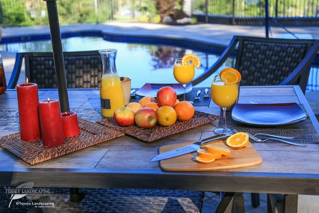 A table with oranges, orange juice, and a knife next to a pool.