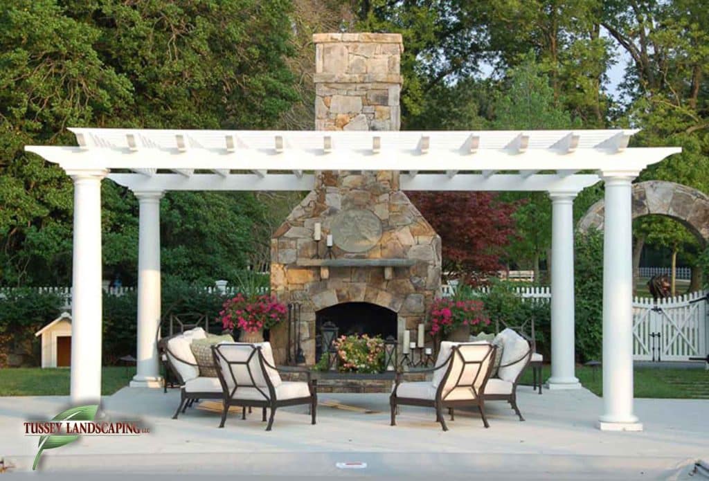 A white pergola with a stone fireplace and patio furniture.