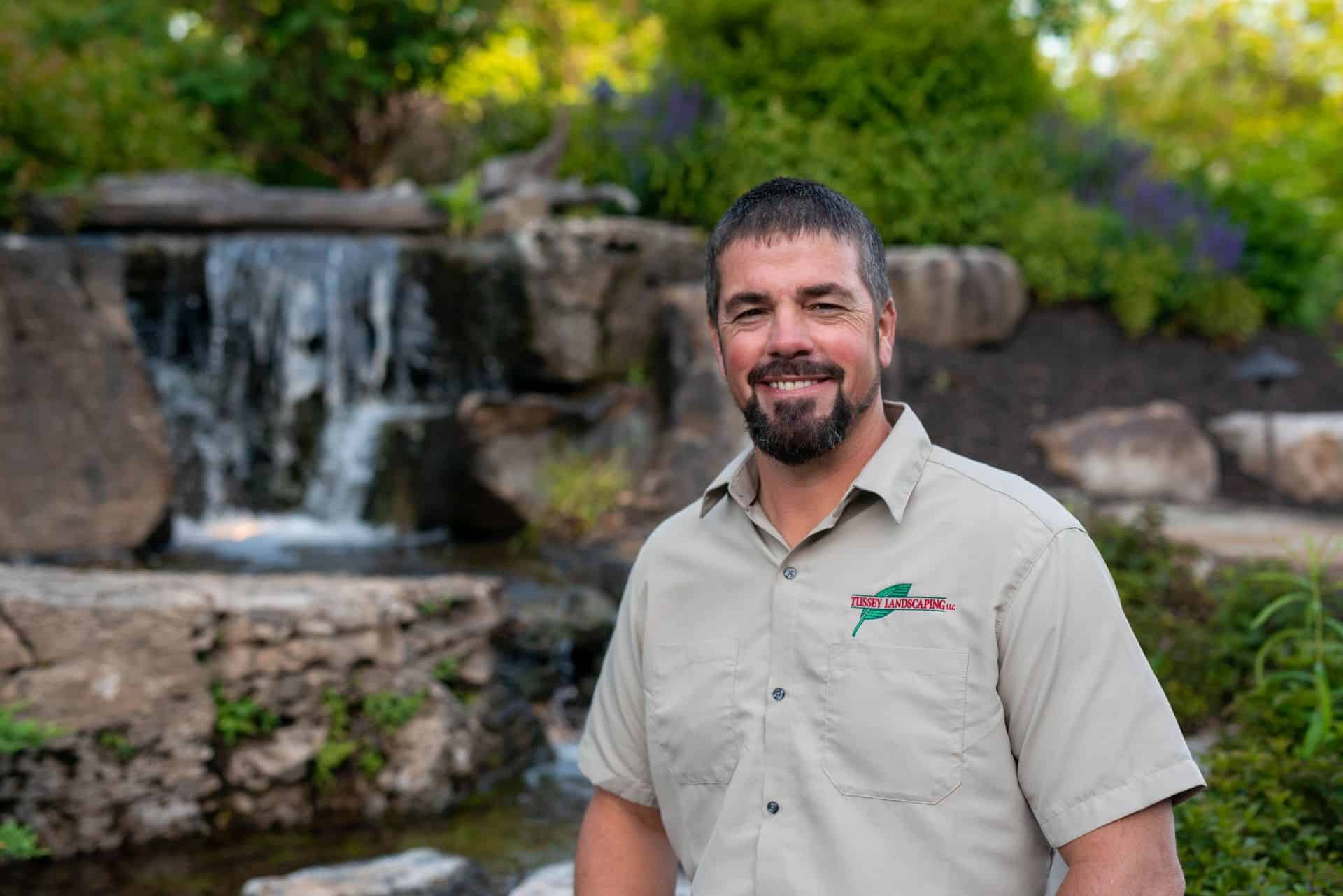 A man in a tan shirt standing in front of a waterfall.