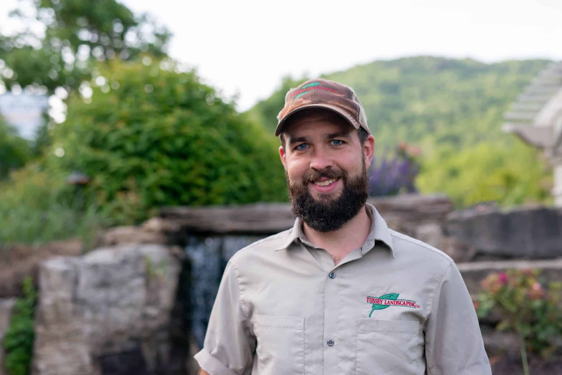 A bearded man in a tan shirt standing in front of a waterfall.