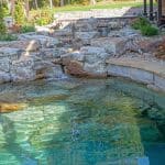swim pond with retaining wall, paver patio, landscaping, water feature, and waterfalls