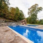 fiberglass swimming pool with paver patio, retaining wall, hot tub, and water slide with landscaping beds
