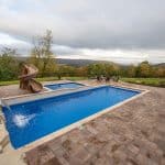 fiberglass swimming pool with paver patio, retaining wall, hot tub, and water slide with landscaping beds