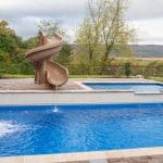 fiberglass swimming pool with paver patio, retaining wall, hot tub, and water slide